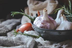 Whole garlic bulbs with a shallot and rosemary sprigs displayed in a rustic metal bowl