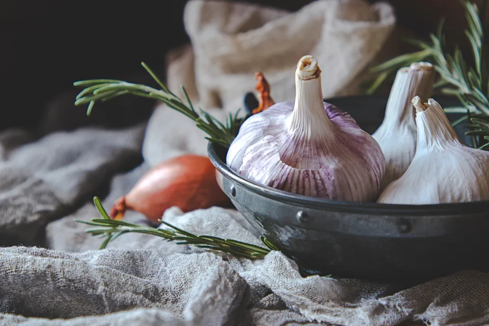 Whole garlic bulbs with a shallot and rosemary sprigs displayed in a rustic metal bowl